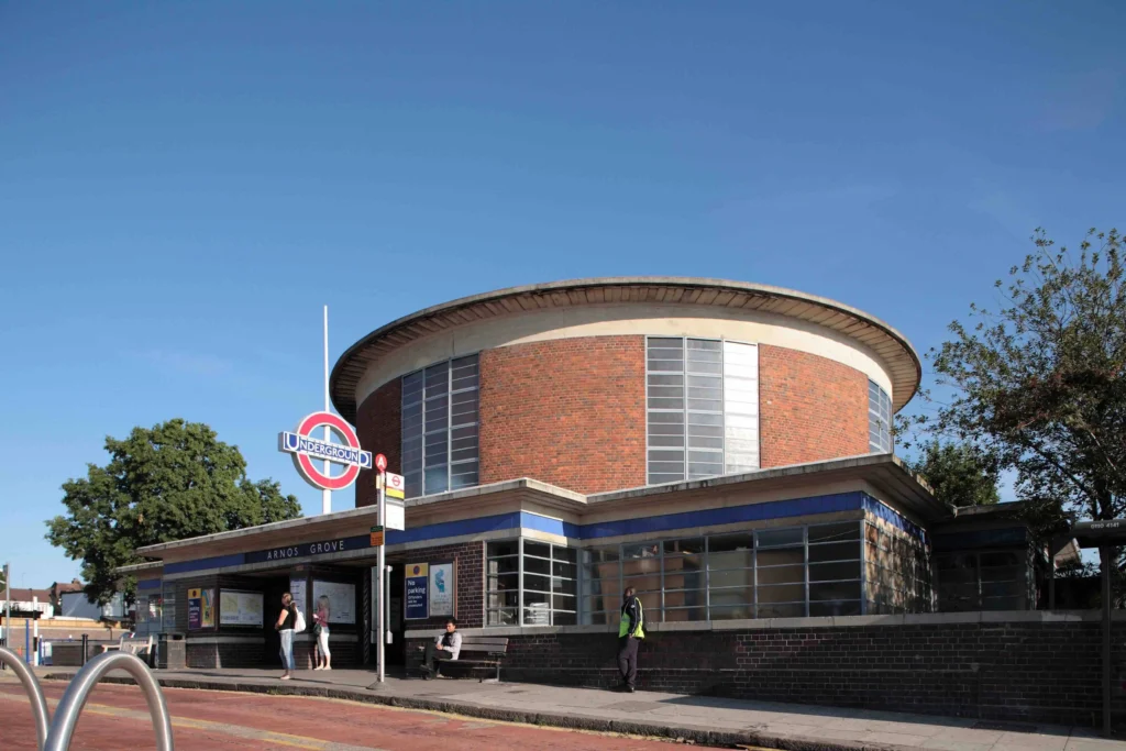 Arnos Grove Station entrance with pedestrians walking past, showing the iconic circular Art Deco building and surrounding North London streets